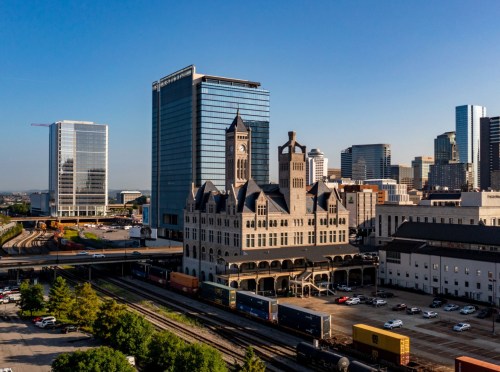 Aerial view of a historic train station surrounded by modern skyscrapers and freight trains in an urban setting.