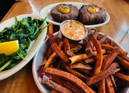 A plate of sweet potato fries, a side of sautéed spinach with lemon, and two beef patties topped with sauce.
