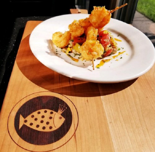 A plate of shrimp skewers on a wooden board with a fish logo, featuring colorful garnishes and a vibrant presentation.