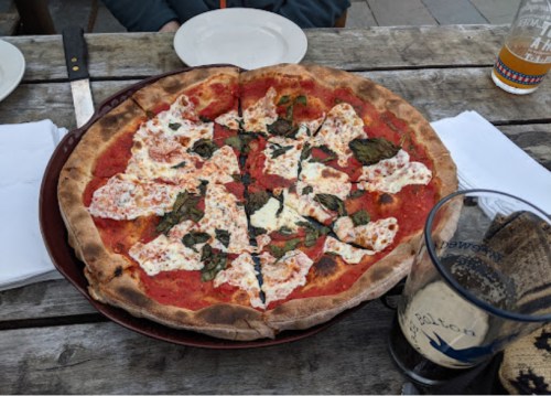 A freshly baked pizza topped with mozzarella, basil, and tomato sauce, served on a wooden table with drinks nearby.