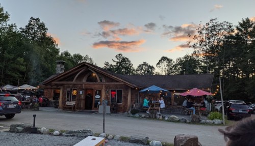 A rustic wooden restaurant with outdoor seating, surrounded by trees and a colorful sunset sky.