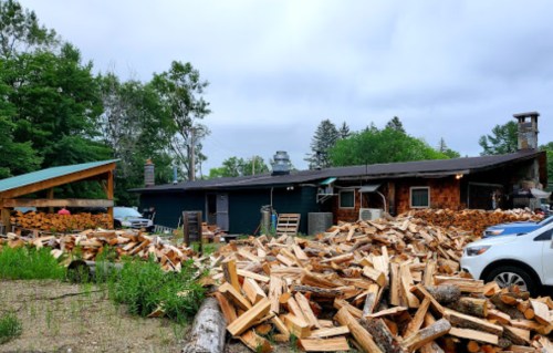 A rustic building surrounded by stacked firewood and greenery under a cloudy sky. A vehicle is parked nearby.