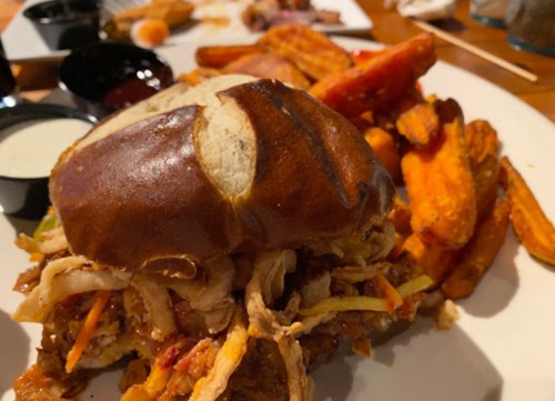 A close-up of a pretzel bun sandwich filled with pulled meat, served with sweet potato fries and dipping sauces.