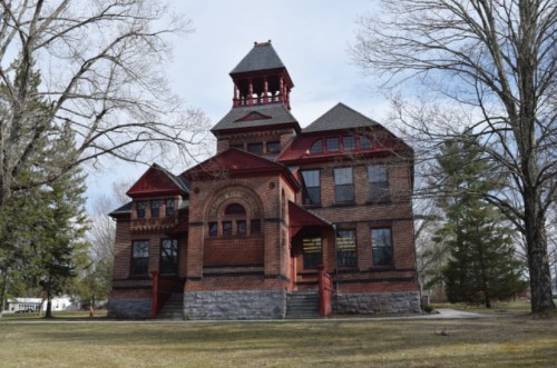 Historic red brick building with a tower, surrounded by trees and a grassy area under a clear blue sky.