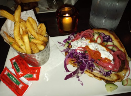 A plate with a pita sandwich filled with vegetables and sauce, alongside a cup of fries and ketchup packets.