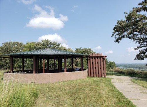 A circular gazebo with a green roof, surrounded by grass and trees, overlooking a scenic landscape under a blue sky.