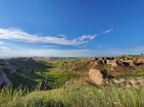 A panoramic view of rolling green hills under a clear blue sky, with rocky formations and sparse trees.