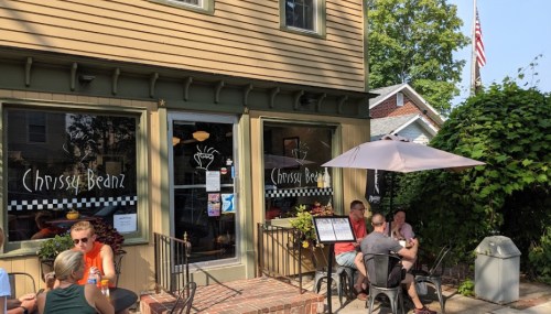A cozy café with outdoor seating, featuring patrons enjoying drinks under an umbrella, surrounded by greenery.