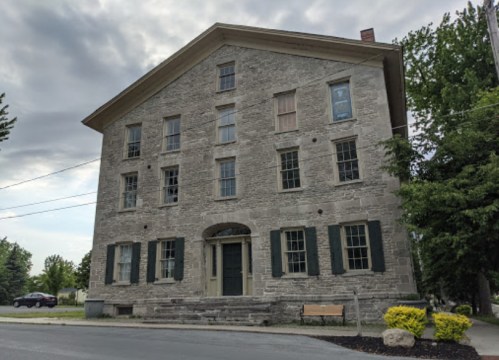 Historic stone building with large windows, green shutters, and a front door, set against a cloudy sky.