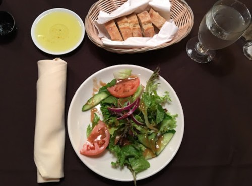 A plate of fresh salad with tomatoes and greens, accompanied by a basket of bread and a small dish of olive oil.
