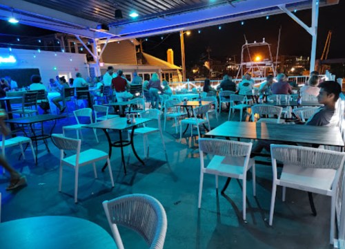 A lively outdoor dining area with tables and chairs, illuminated by blue lights, and people enjoying the atmosphere.