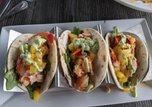 Three tacos filled with shrimp, mango, and avocado sauce, served in a metal holder on a white plate.
