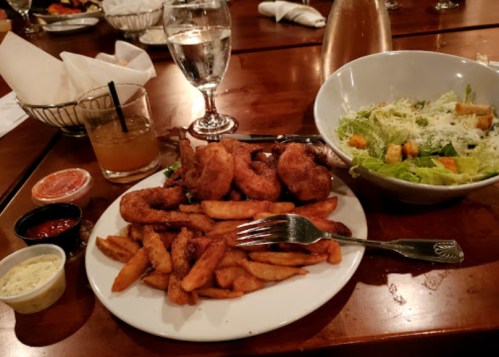 A plate of fried shrimp and fries, accompanied by a Caesar salad and drinks on a wooden table.