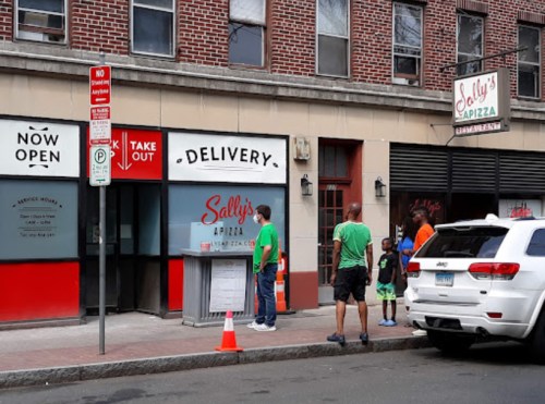 People stand outside Sally's Pizza, which has a "Now Open" sign, with a delivery and takeout window visible.