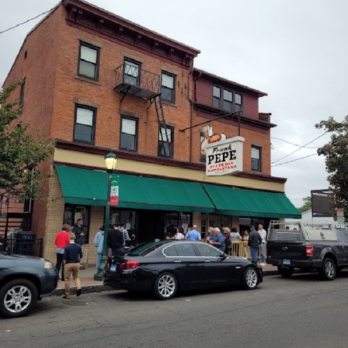 A busy pizzeria with a green awning, people waiting outside, and parked cars in front of a brick building.