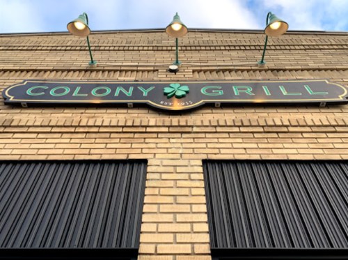 Sign for Colony Grill, featuring a green clover, mounted on a brick wall with lights above.