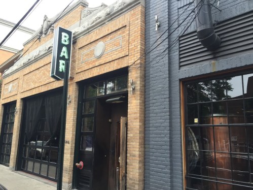 Exterior of a brick building featuring a bar sign, large windows, and a rustic entrance.