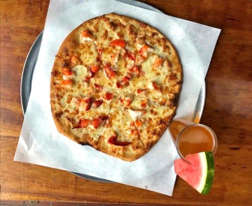 A freshly baked pizza topped with red peppers and cheese, served on a metal tray with a glass of drink and watermelon slice.