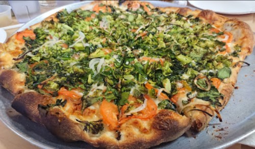 A large pizza topped with fresh vegetables, including broccoli, tomatoes, and herbs, on a metal tray.