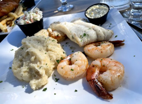 A plate featuring grilled fish, shrimp, creamy mashed potatoes, and a side of coleslaw with dipping sauce.
