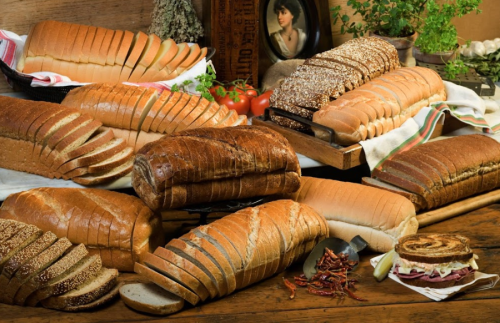 A variety of sliced bread loaves arranged on a wooden table, with tomatoes and herbs in the background.