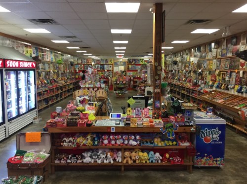 A colorful candy store filled with shelves of sweets, toys, and snacks, featuring a soda fridge and a vibrant checkout area.