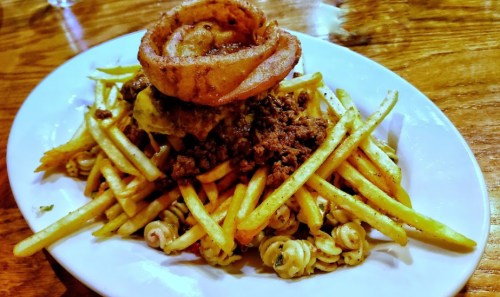 A plate of pasta topped with seasoned ground meat, French fries, and a crispy onion ring.