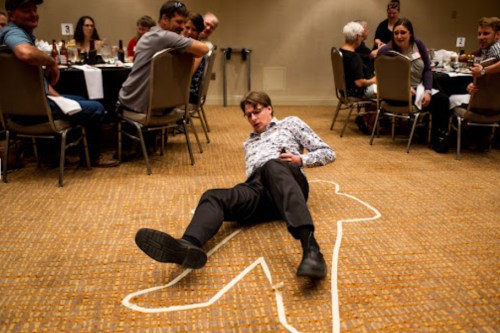 A man lies on the floor in a chalk outline, while amused guests watch from nearby tables at a dinner event.