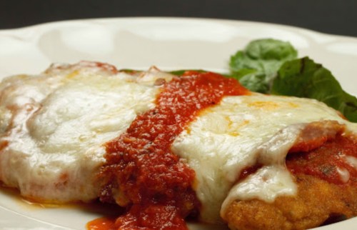 A close-up of a cheesy chicken parmesan topped with marinara sauce and a side of fresh greens on a white plate.