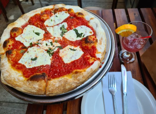 A freshly baked pizza with mozzarella and basil on a wooden table, accompanied by a cocktail and plates.