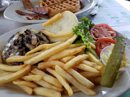 A plate with a grilled burger topped with mushrooms, fries, lettuce, tomato, pickle, and a slice of bread.