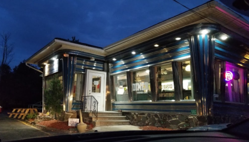 A brightly lit diner with a blue exterior, featuring large windows and a welcoming entrance at dusk.