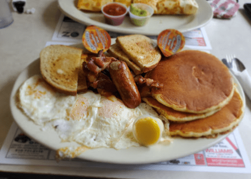 A breakfast plate featuring pancakes, eggs, bacon, sausage, and toast, with syrup on the side.