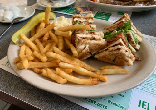 A plate with a club sandwich, lettuce, tomato, and fries, accompanied by a pickle and a small dish of butter.