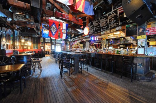 A spacious bar interior with wooden floors, a long counter, and various flags hanging from the ceiling.