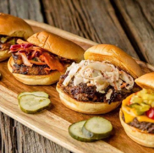 Three gourmet burgers on a wooden platter, each topped with different ingredients, alongside slices of pickles.