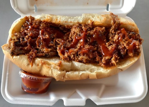 A pulled pork sandwich topped with barbecue sauce, served in a foam container with a side of sauce.