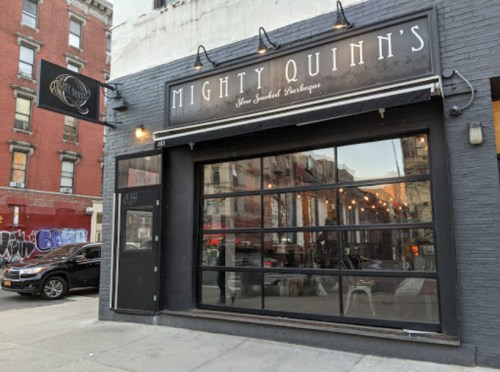 Exterior of Mighty Quinn's, a barbecue restaurant, featuring large glass windows and a sign above the entrance.