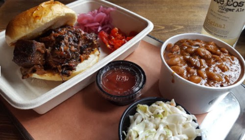 A pulled pork sandwich with pickled onions, tomatoes, baked beans, and coleslaw in a takeout container.