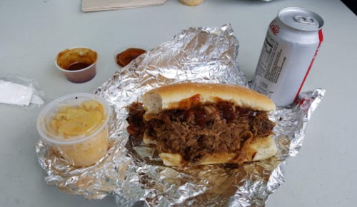 A foil-wrapped sandwich filled with shredded meat, a side of sauce, and a can of soda on a table.