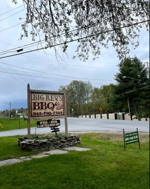 Sign for Big Kev's BBQ with contact information, surrounded by greenery and a cloudy sky. Road visible in the background.