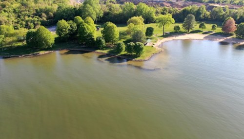 Aerial view of a serene lake surrounded by lush green trees and a sandy shore on a sunny day.