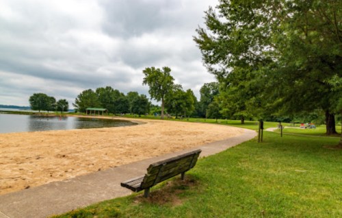 A peaceful lakeside scene featuring a sandy beach, a bench, and lush greenery under a cloudy sky.