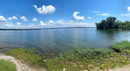 A panoramic view of a calm lake surrounded by greenery and blue skies with fluffy clouds.