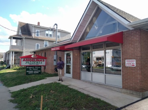 A person stands outside Angelina's Pizzeria, a brick building with a red awning and signage, on a sunny day.