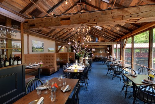 Cozy restaurant interior with wooden beams, elegant tables, and large windows overlooking greenery.
