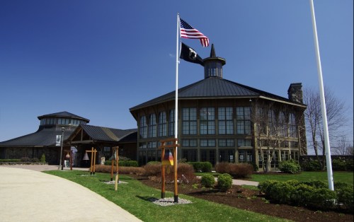 A modern building with large windows, surrounded by greenery, featuring American and POW flags in front.