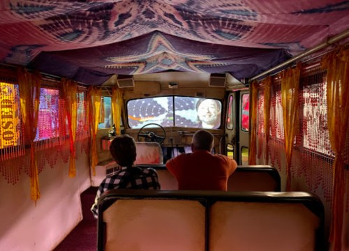 Two people sit inside a colorful, retro camper van, watching a projection on the front window. Vibrant decor surrounds them.