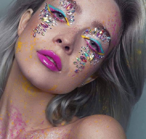 A close-up of a woman with colorful makeup, sparkling gems on her face, and vibrant pink lips against a soft background.