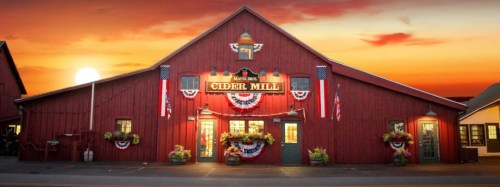 A red barn-style cider mill adorned with flags and bunting, set against a sunset sky.